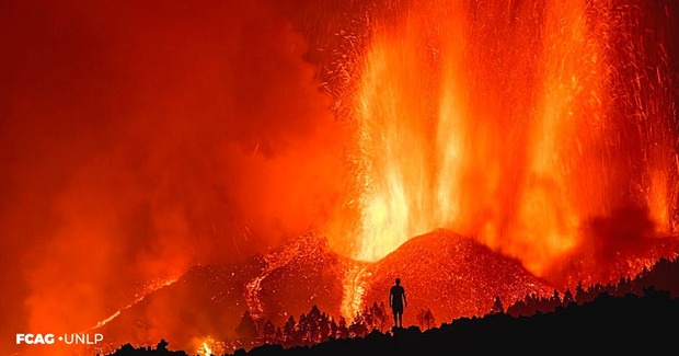 La imagen muestra la erupción  en colores rojizos y anaranjados y la lava que cae por las laderas. Se observa el contorno de una persona observando la escena.